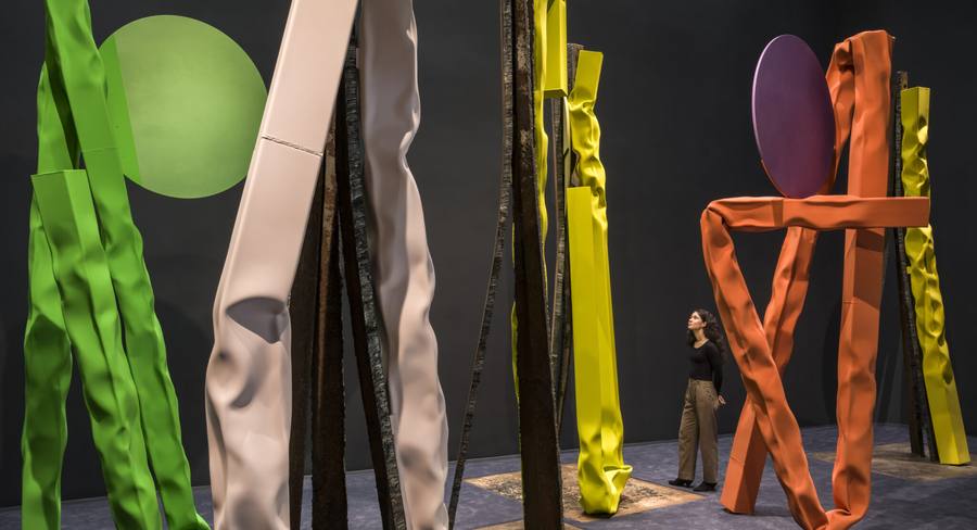 Installation view of Carol Bove at the Guggenheim, with monumental crumpled steel sculptures in green, pink, yellow and orange and a visitor standing among them for scale