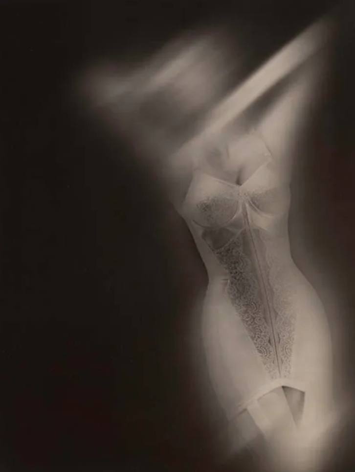 Black and white fashion photograph by Lillian Bassman of a woman in a pale lace corselette, high in contrast and softly blurred so that the garment dissolves into light against a dark ground