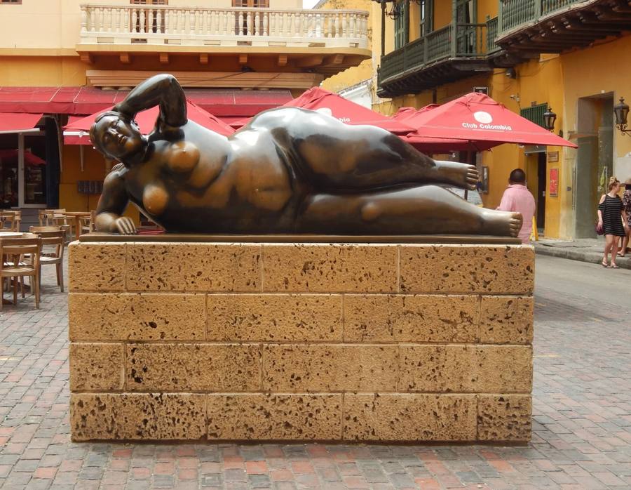 Fernando Botero's monumental bronze sculpture of a full bodied woman reclining on her side, on a stone plinth in a square in Cartagena, with a yellow colonial building and red cafe umbrellas behind it