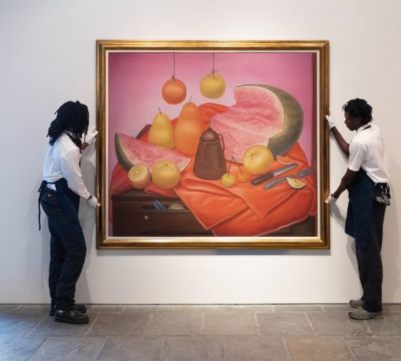 Fernando Botero still life painting of a sliced watermelon, oranges, pears and other fruit arranged on an orange cloth against a pink background, held on a gallery wall by two white gloved handlers