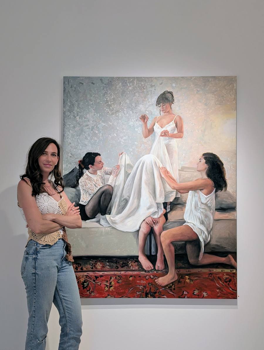 Curator Stav Theodor Kimhi standing beside a large Tom Fima painting of three young women in white sewing and mending fabric on a bed, at Gallery Be'eri in Tel Aviv