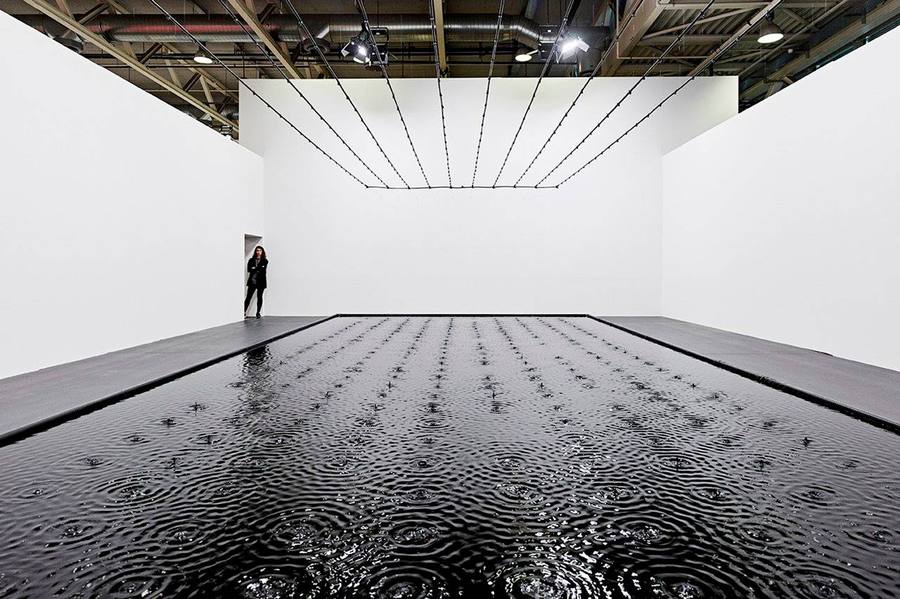 Belu-Simion Fainaru installation: a large rectangular black pool set into a white gallery floor, a grid of drippers suspended from the ceiling releasing drops that ripple across the dark water, a single figure standing at the far wall for scale
