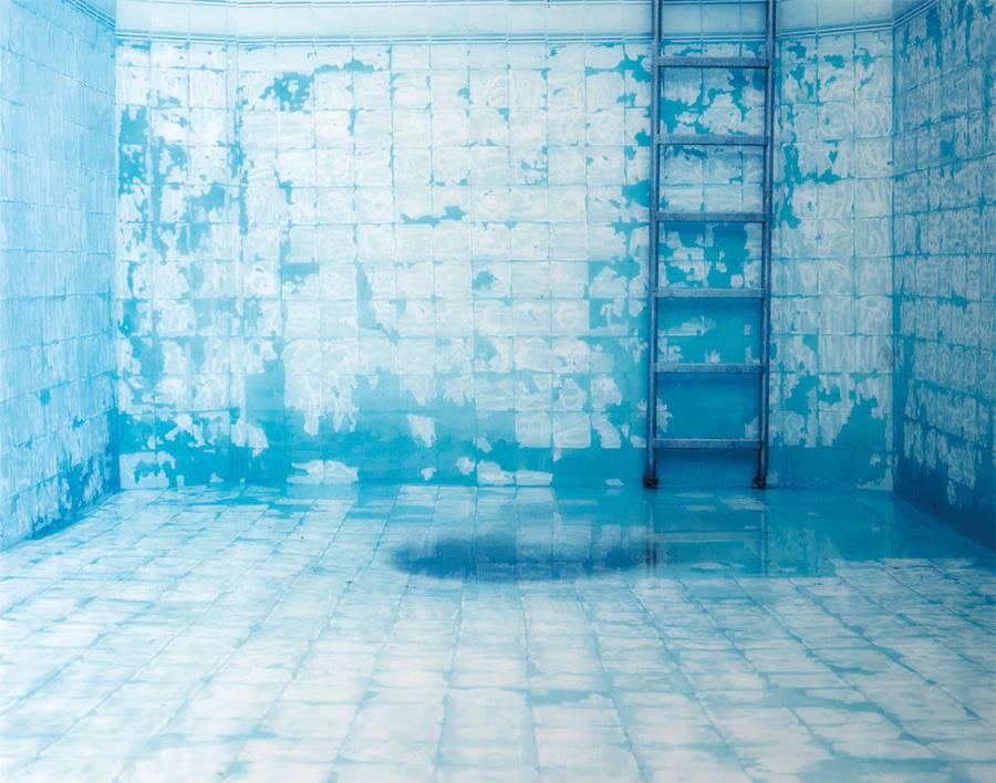 Tal Shochat photograph of an empty, drained swimming pool interior tiled entirely in pale blue, the tiles worn and mottled with white, a metal ladder against the far wall, no water and no people