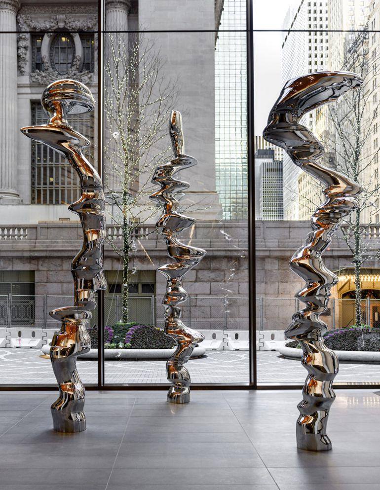 Three tall polished stainless steel Tony Cragg sculptures with twisting stacked forms standing in the glass-walled lobby of One Vanderbilt, Grand Central Terminal visible through the windows behind them
