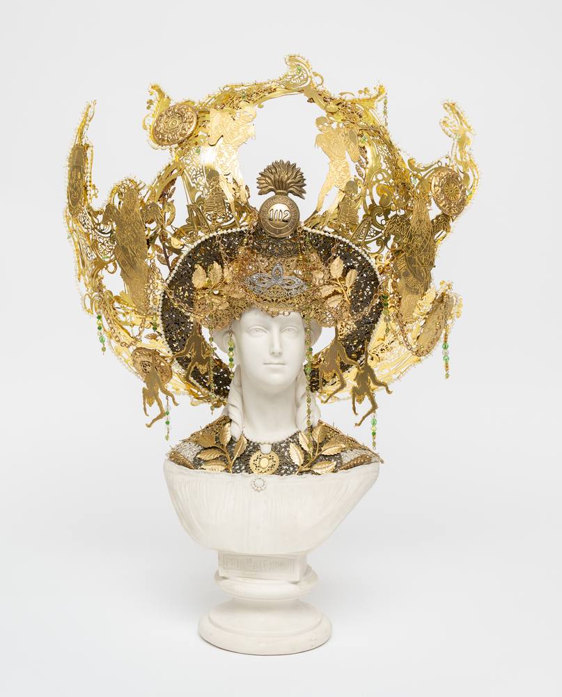 Hew Locke sculpture, a white Parian ware bust of a woman crowned with an elaborate radiating headdress of gold cut metal, filigree, beads and small hanging green stones, a numbered medal reading 1002 at the center