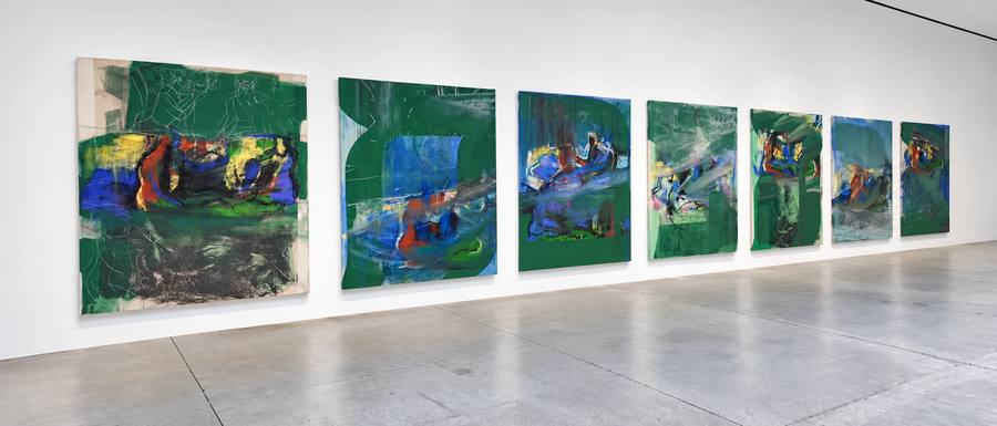 Installation view of seven large Rita Ackermann paintings hung in a row on a long white gallery wall above a polished concrete floor, each canvas dominated by deep green chalkboard paint with bursts of blue, red and yellow