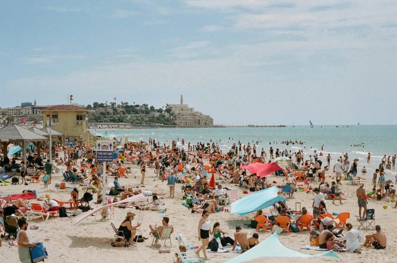 Analog photograph of a crowded Tel Aviv beach in daylight, with sunbathers, orange beach chairs and colorful shade cloths on the sand, the sea beyond and the old city of Jaffa on the headland
