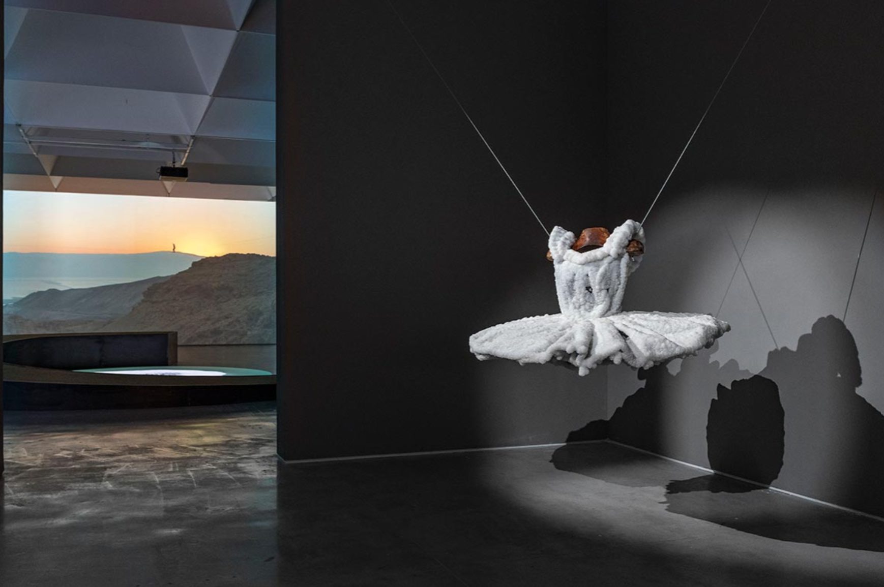 Installation view of Sigalit Landau's white ballet tutu, crusted with Dead Sea salt crystals and hung by two wires in a dark gallery corner, with a desert sunrise video projected in the next room