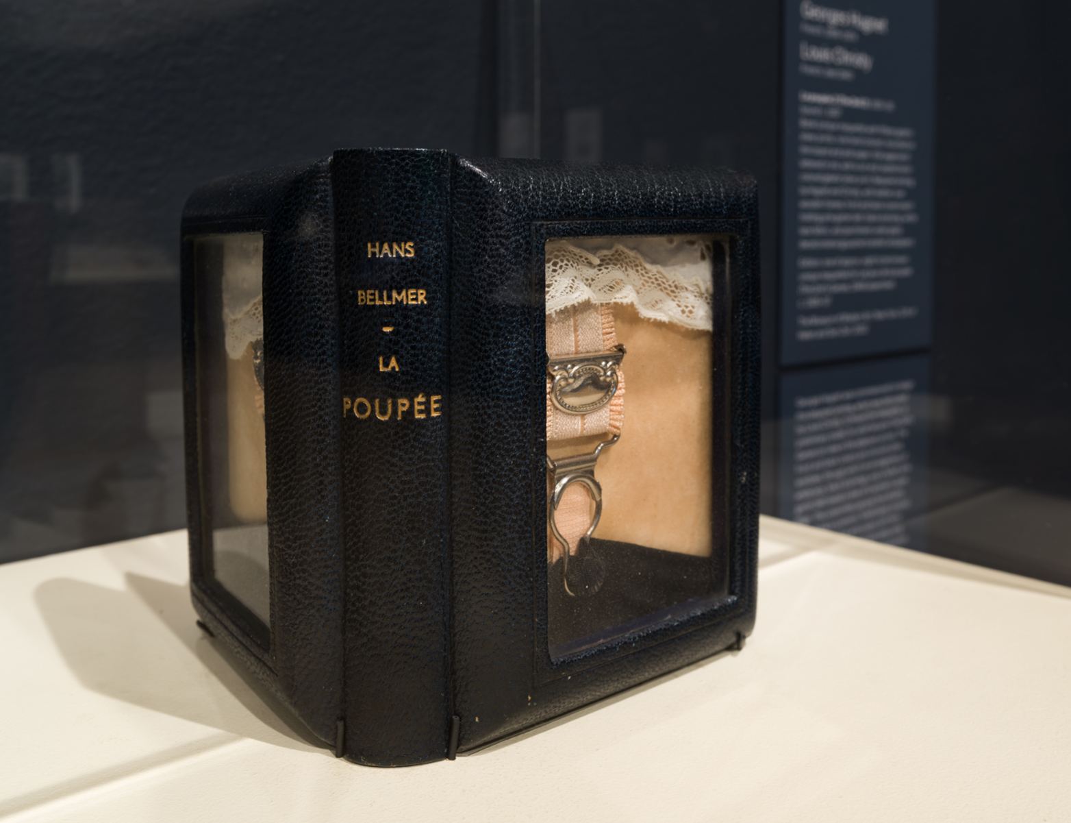 A black leather bound book object with a gilt spine reading Hans Bellmer, La Poupee, fitted with glass windows through which a lace edged garter can be seen, in a museum vitrine