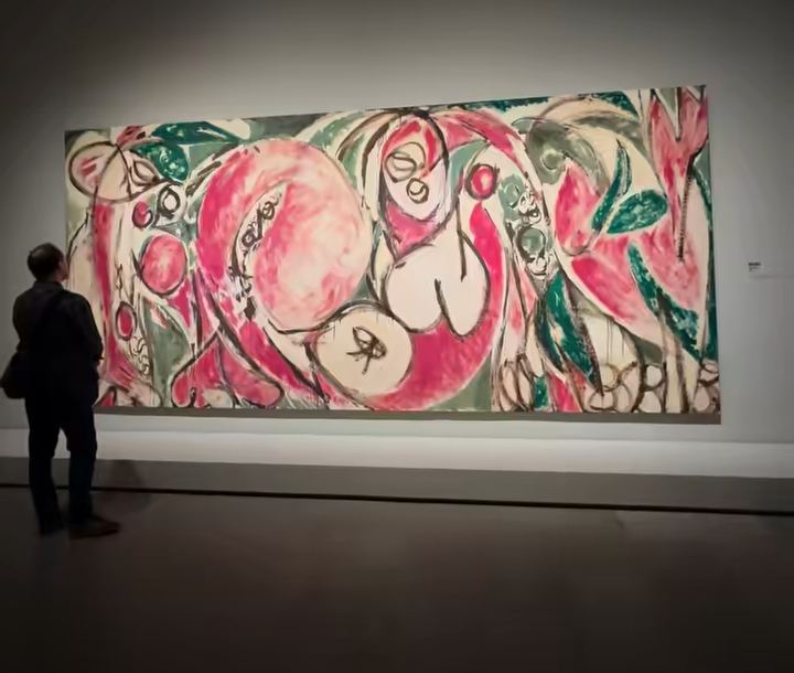 A visitor in dark clothing stands before a very large Lee Krasner painting on a gallery wall, with rounded pink, green and cream forms outlined in black