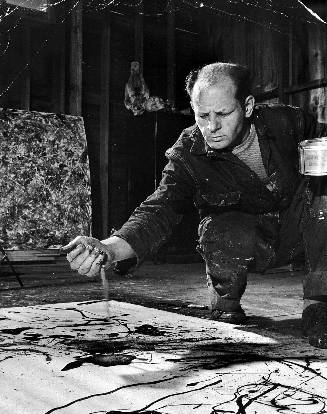 Black and white photograph of Jackson Pollock crouching in his barn studio, letting a stream of sand or paint fall from his hand onto a canvas laid on the floor, with a finished drip painting leaning on the wall behind him