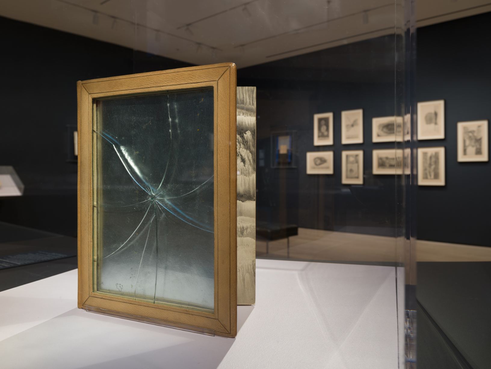 A book with a tan leather cover framing a sheet of cracked glass, standing open on a white plinth in a dark gallery with framed prints on the far wall