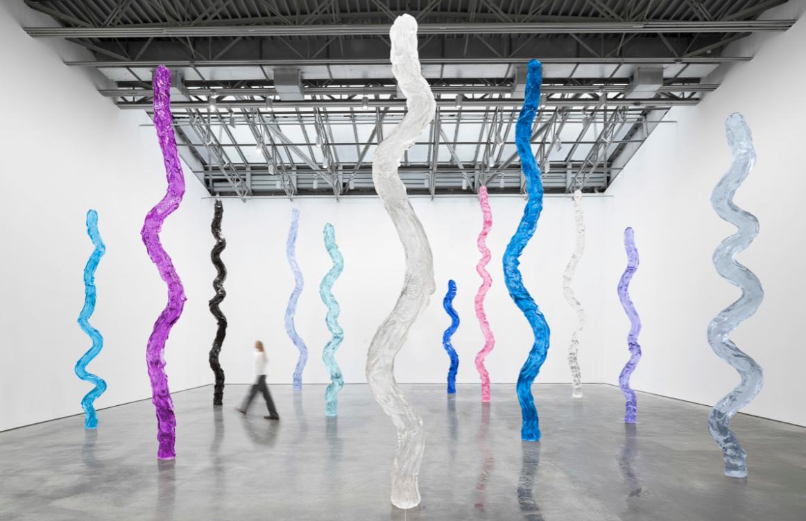 Installation view of Ugo Rondinone's waterfall: a dozen tall, undulating translucent sculptures in purple, blue, pink, black, white and pale tints standing in a large white gallery, with a visitor walking among them