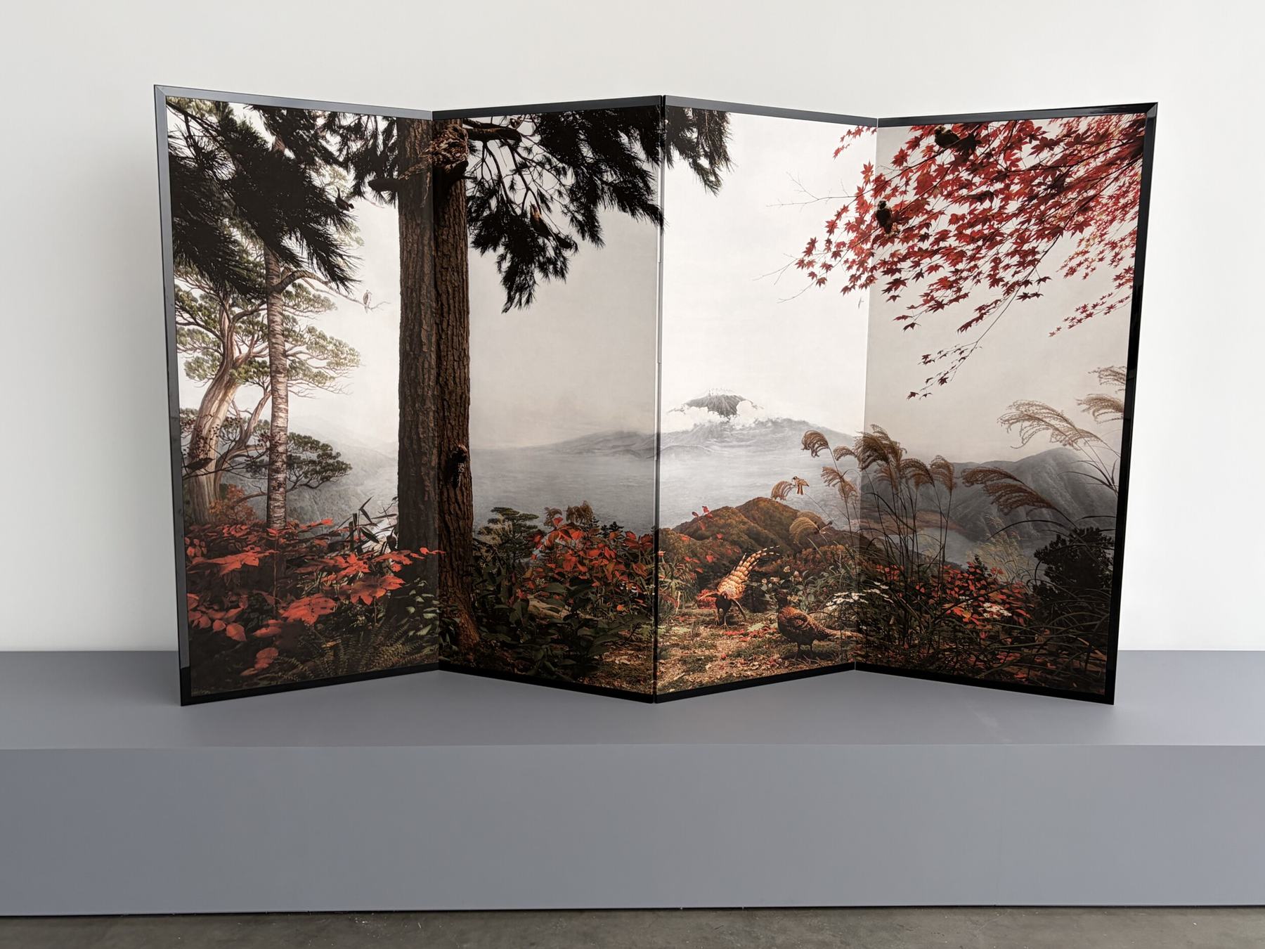 A four panel byobu folding screen printed with a color photograph of a diorama of an autumn mountain forest, with pine trees, red maple leaves, pampas grass, pheasants and a distant snow capped peak, standing on a gray plinth in a white gallery