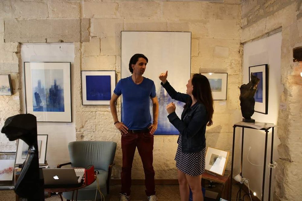 Stav Theodor in a denim jacket talking with an artist in a stone walled gallery hung with blue canvases