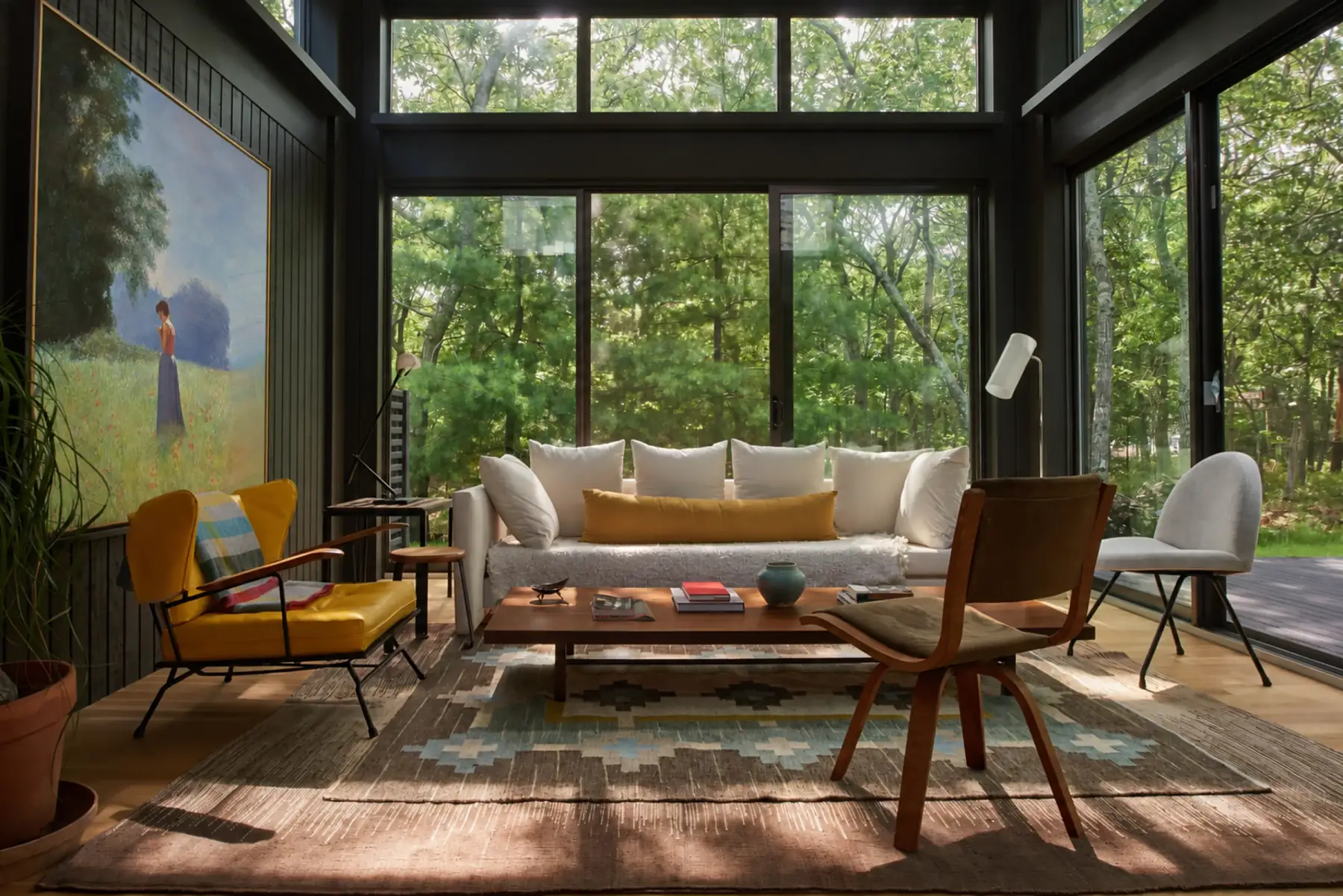 A glass-walled living room in the trees with a tall painting of a girl in a meadow on the left wall.