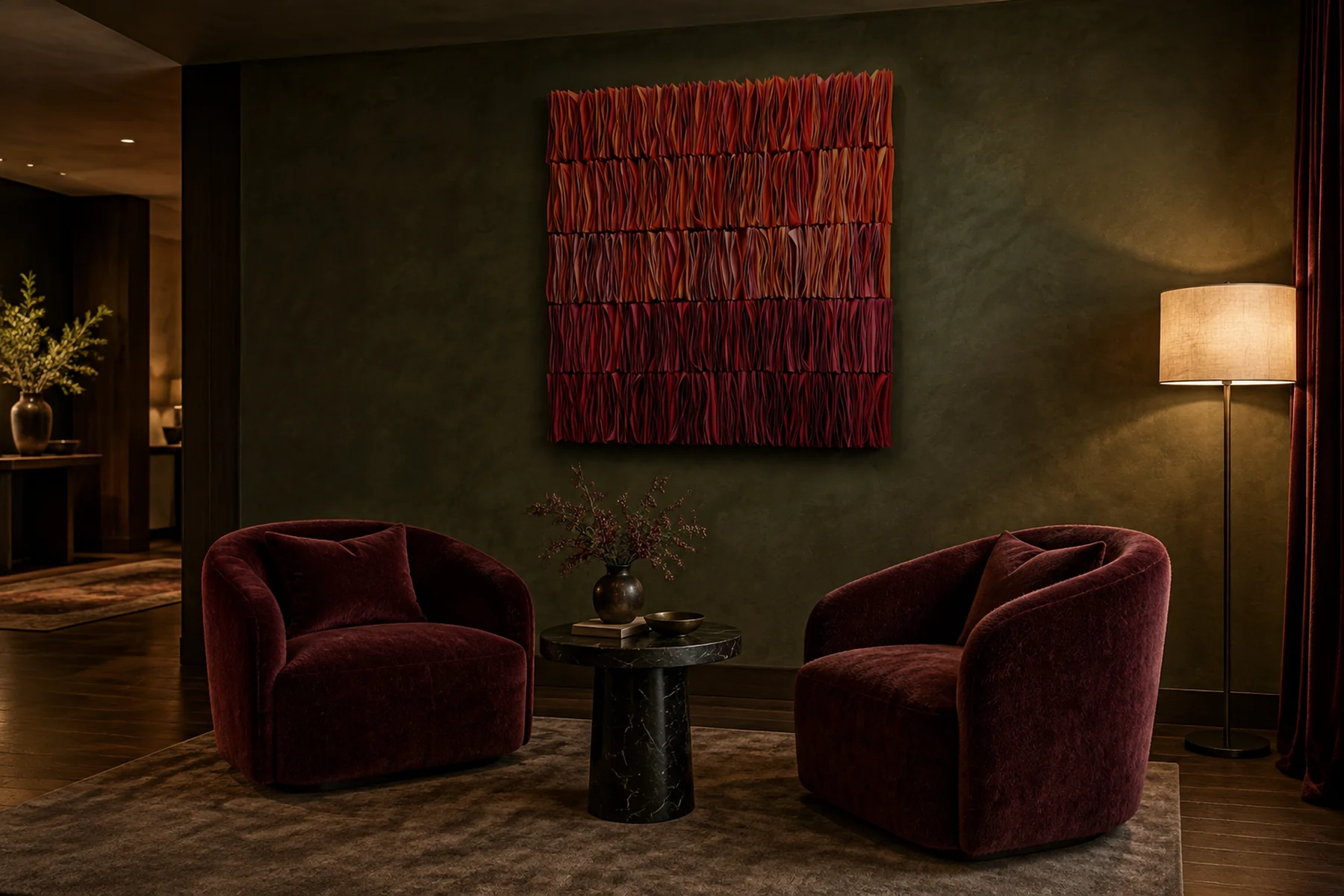 A dim hotel lounge: a tall wall work of folded strips in reds from coral to burgundy on an olive plaster wall above two velvet chairs.