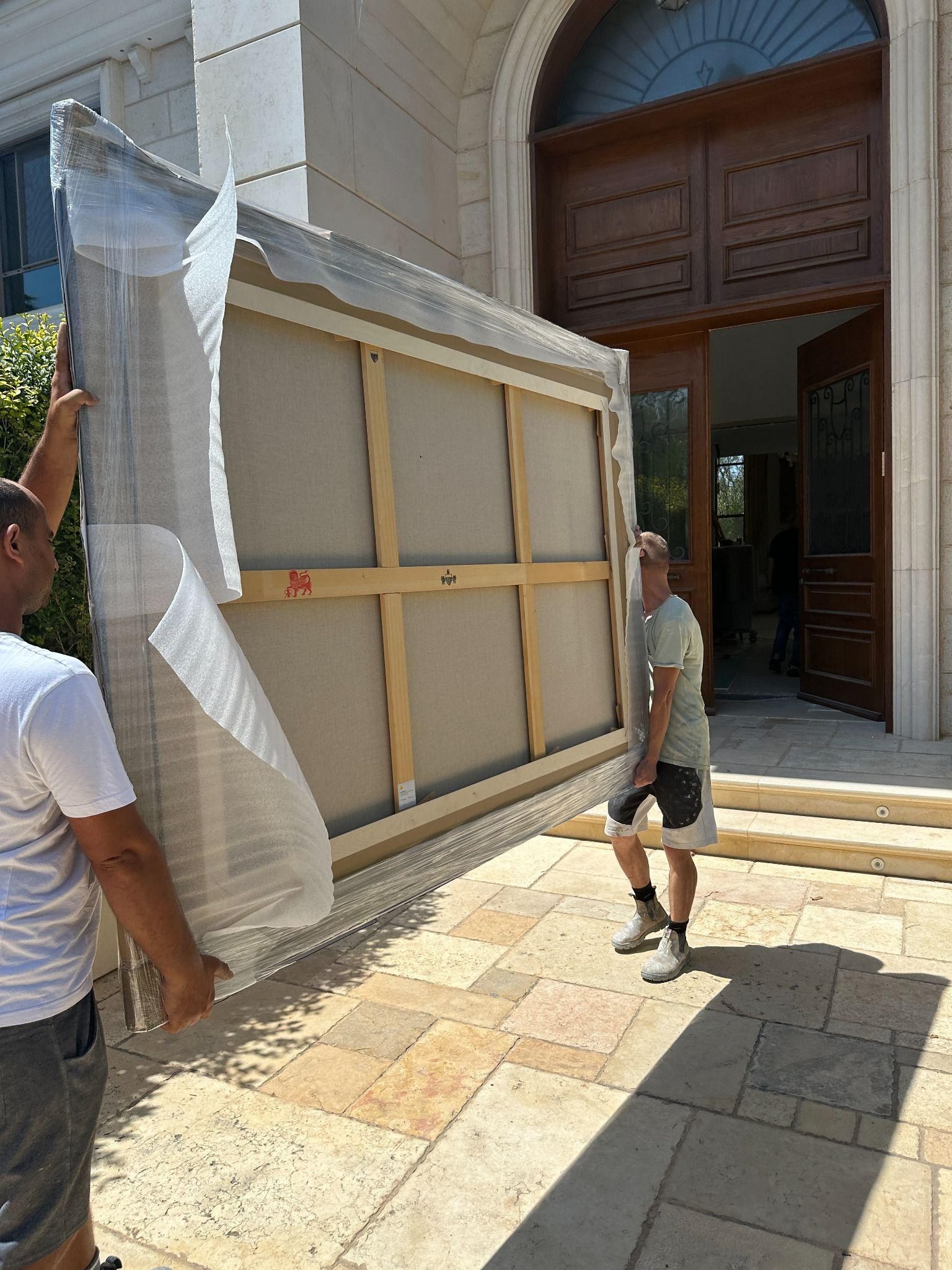 Two art handlers carrying a large wrapped canvas through the entrance of a private estate in Caesarea