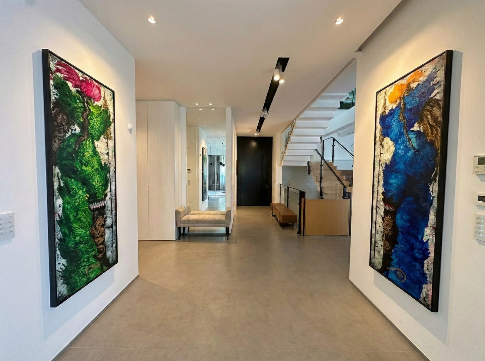 Foyer of a Caesarea garden villa: two tall portraits in slim black frames, one in greens with a pink headdress, one in blues with orange hair, on the walls of the entrance hall, black front door and staircase beyond