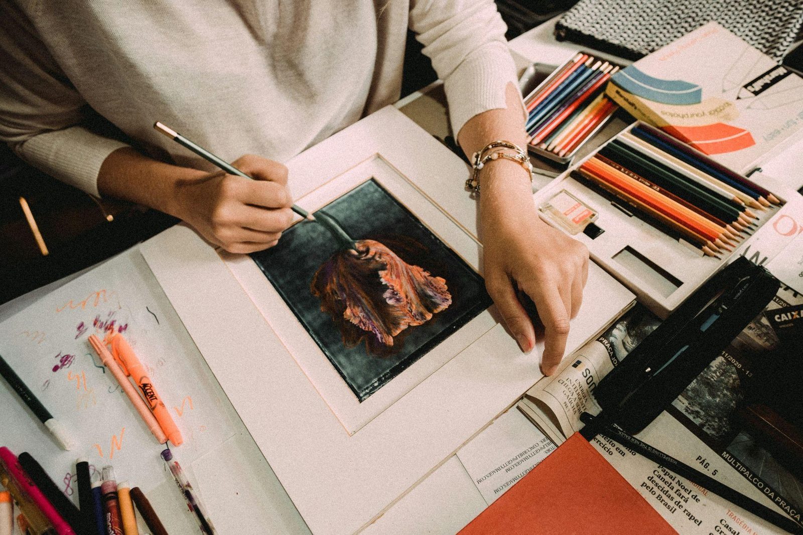 A framer's table: hands positioning a small dark floral painting inside a white mat, with pencils and framing tools around it